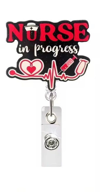 A nurse-themed retractable badge reel with a black and red 'NURSE in progress' design, white script text, and medical icons including a heart monitor line, heart, syringe, and stethoscope. The reel body is white with a silver metal swivel and ring hardware.